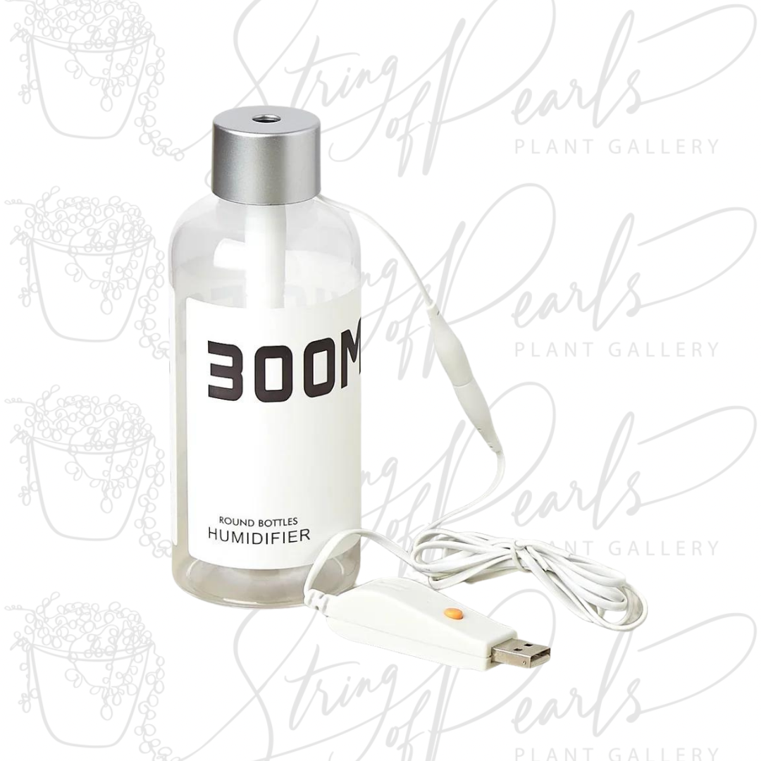 A clear bottle-style humidifier labeled "300ML" with a silver cap and a USB-powered cord attached. The humidifier is designed for adding moisture to the air in small spaces, making it suitable for indoor plant care or personal use. The simple, compact design allows for easy portability and use in various settings.